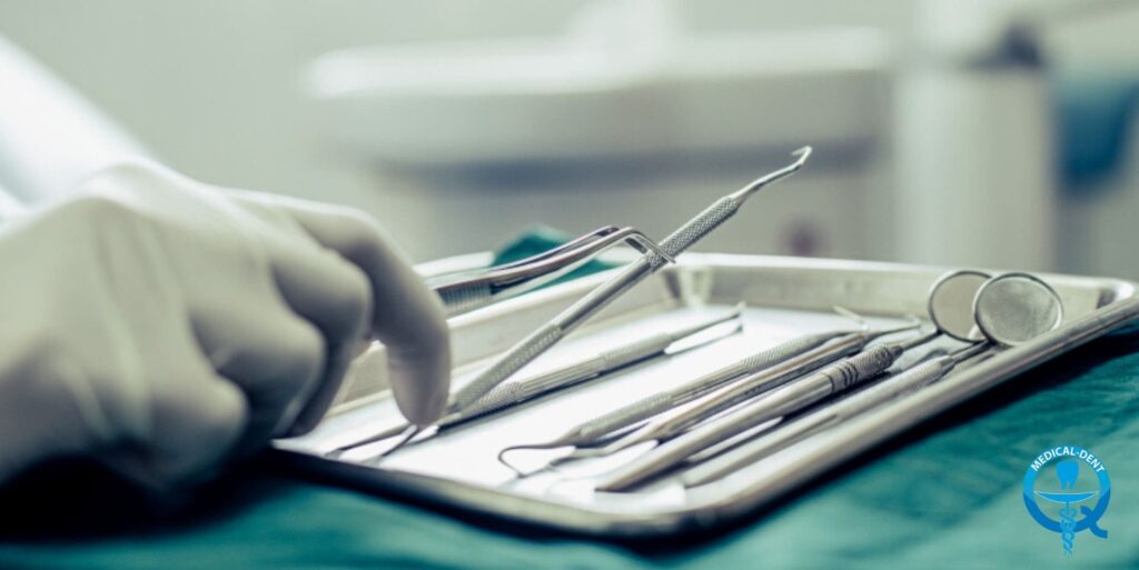 Zdjęcie przedstawia zestaw sterylnych narzędzi dentystycznych na metalowej tackce. Widoczne są szczypce, zgłębniki, skalpele i inne drobne instrumenty wykorzystywane w zabiegach stomatologicznych, takich jak pokrycie recesji dziąsłowej. Ręka w niebieskiej rękawiczce trzyma niektóre z narzędzi, prawdopodobnie przygotowując się do zabiegu.