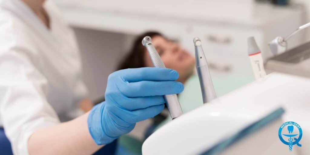 Obraz przedstawia zbliżenie na dłonie dentysty trzymające narzędzia stomatologiczne podczas zabiegu. Na drugim planie widoczna jest rozmyta twarz osoby poddawanej zabiegowi. Zdjęcie ilustruje temat zgorzelizbu i leczenia zębów.