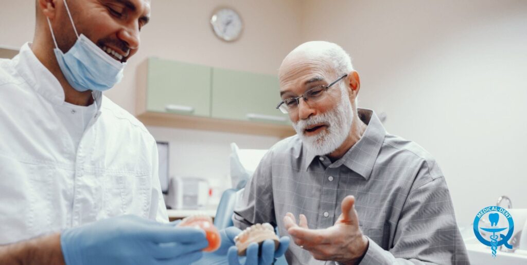 Obraz przedstawia starszego mężczyznę z siwą brodą, noszącego okulary i siedzącego na krześle podczas wizyty u dentysty. Dentysta w niebieskim fartuchu i masce medycznej przeprowadza konsultację z pacjentem, wskazując na jego uzębienie. Scena rozgrywa się w gabinecie stomatologicznym, co sugeruje, że omawiana jest kwestia protez zębowych.