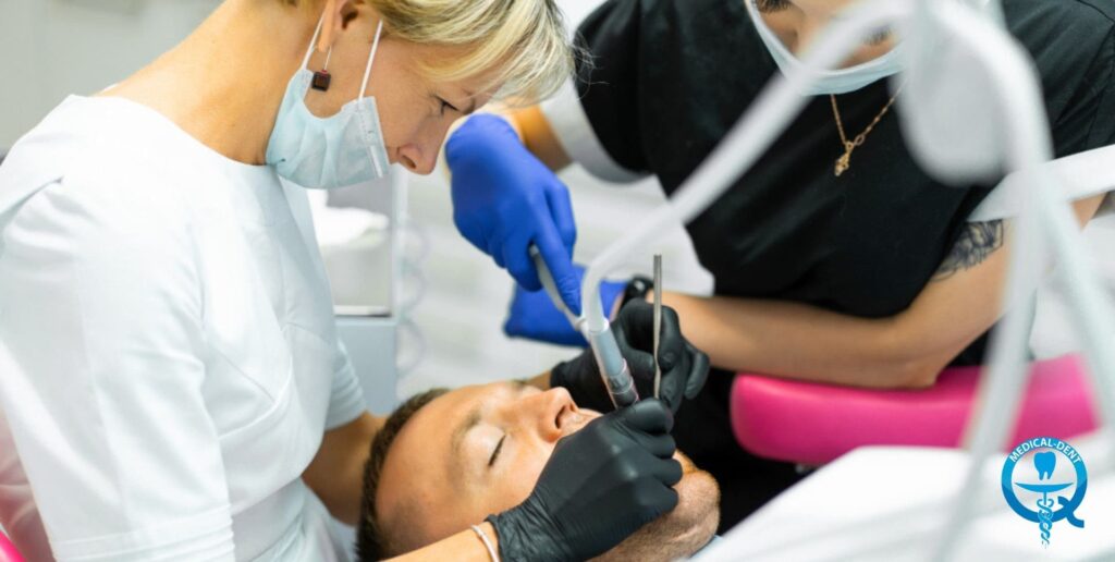 Obraz przedstawia zabieg stomatologiczny, podczas którego dwie osoby w fartuchach medycznych i maskach wykonują procedurę na pacjencie leżącym na fotelu dentystycznym. Jedna z osób, prawdopodobnie dentystka, trzyma w dłoni niebieski narzędzie dentystyczne, podczas gdy druga asystuje przy zabiegu. Zabieg wydaje się dotyczyć korzenia zęba pacjenta.