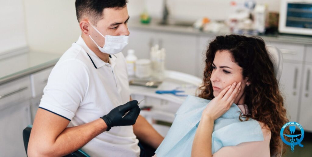 Zdjęcie przedstawia pacjentkę siedzącą na fotelu stomatologicznym i przykładającą dłoń do ust, podczas gdy dentysta w masce i rękawiczkach bada jej zęby. Obraz ilustruje temat przodozgryzu i jego leczenia. Osoby nie są rozpoznawalne.