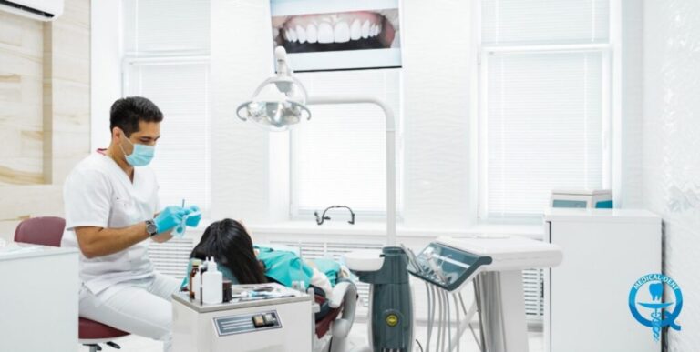 Obraz przedstawia dentystę wykonującego zabieg stomatologiczny na pacjencie. Widoczne są urządzenia i narzędzia obecne w gabinecie stomatologicznym, takie jak unit stomatologiczny, lampa operacyjna i monitor do wyświetlania zdjęć zębów. Zarówno dentysta, jak i pacjent noszą maseczki ochronne.