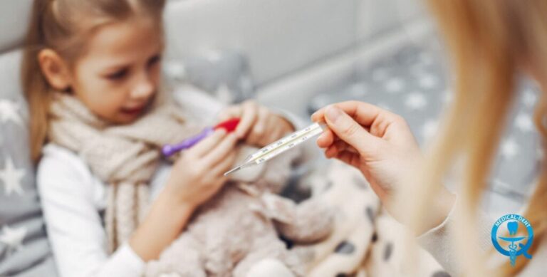 Obraz przedstawia małą dziewczynkę siedzącą na łóżku. Jej twarz ma lekko zmarkowany wyraz, sugerujący że odczuwa ból. Obok niej znajduje się osoba dorosła, trzymająca w dłoni termometr bezdotykowy. Sytuacja sugeruje, że sprawdzana jest temperatura dziecka, prawdopodobnie w związku z przeziębieniem lub bólem zęba.