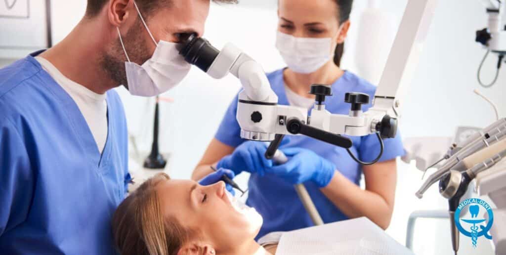 Alt text: "Dentysta w niebieskim fartuchu używa mikroskopu stomatologicznego podczas zabiegu odsłonięcia zęba zatrzymanego. Asystentka w masce i niebieskim uniformie pomaga przy zabiegu. Pacjent leży na fotelu dentystycznym. W tle widoczny jest sprzęt stomatologiczny."