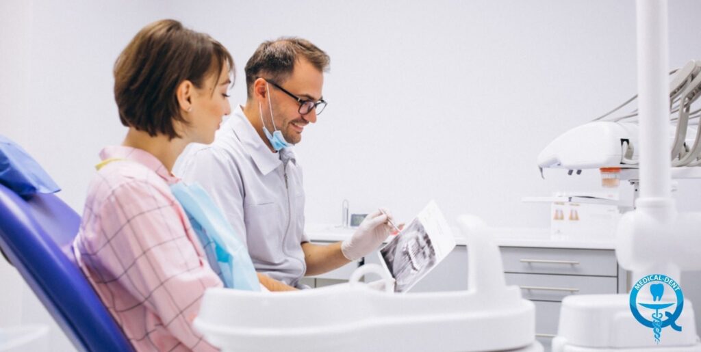 Alt text: "W nowoczesnym gabinecie stomatologicznym dentysta w białym fartuchu i masce konsultuje zdjęcie rentgenowskie z pacjentką siedzącą na fotelu dentystycznym. W tle widoczny jest sprzęt stomatologiczny i logo kliniki."