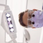 "Widok z perspektywy pacjenta na dentystę w masce ochronnej, okularach i niebieskim fartuchu, pochylającego się nad fotelem dentystycznym. W kadrze widoczna jest również dentystyczna lampa zabiegowa z białą obudową i jasnymi diodami LED."