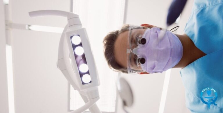 "Widok z perspektywy pacjenta na dentystę w masce ochronnej, okularach i niebieskim fartuchu, pochylającego się nad fotelem dentystycznym. W kadrze widoczna jest również dentystyczna lampa zabiegowa z białą obudową i jasnymi diodami LED."