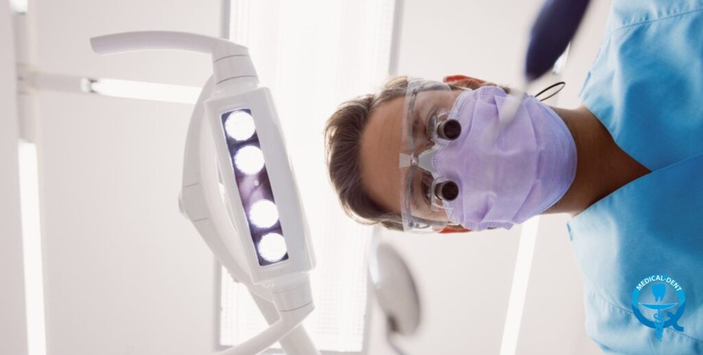 "Widok z perspektywy pacjenta na dentystę w masce ochronnej, okularach i niebieskim fartuchu, pochylającego się nad fotelem dentystycznym. W kadrze widoczna jest również dentystyczna lampa zabiegowa z białą obudową i jasnymi diodami LED."