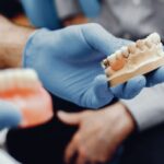 Gloved hands holding dental prosthetic models, including a partial denture, in a clinical setting.