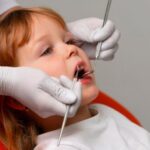 Dziecko leżące na fotelu dentystycznym, podczas gdy dentysta w białych rękawiczkach używa narzędzi do badania jego jamy ustnej.