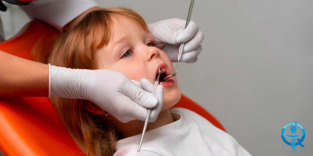 "Dziecko podczas badania stomatologicznego, leżące na pomarańczowym fotelu dentystycznym. Dentysta w białych rękawiczkach wykonuje przegląd jamy ustnej przy użyciu standardowych narzędzi. W prawym dolnym rogu widoczne niebieskie logo gabinetu stomatologicznego."