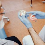 Dentysta w niebieskich rękawiczkach prezentuje modele protez pacjentowi z białą brodą i okularami w gabinecie stomatologicznym.