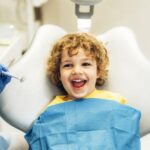 Dziecko na fotelu dentystycznym, dentysta w rękawiczkach trzyma narzędzie do fluoryzacji, dziecko ma uśmiech.