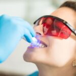 Zabieg wybielania zębów: dentysta w rękawiczkach aplikuje preparat na zęby pacjenta za pomocą urządzenia, pacjent nosi ochronne okulary.