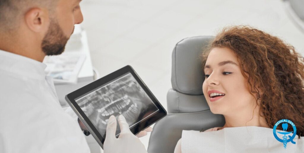 Dentysta w białym fartuchu i rękawiczkach pokazuje pacjentce w fotelu dentystycznym zdjęcie rentgenowskie zębów na tablecie.