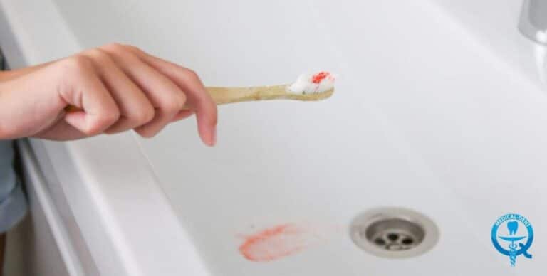 Ręka trzyma szczoteczkę do zębów z pastą i krwią na włosiu, nad umywalką z plamą krwi w odpływie.