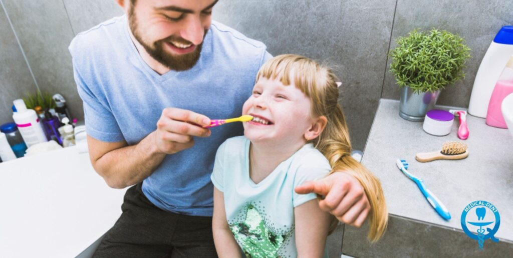 Adult male helping a young girl brush her teeth in a bathroom with toiletries and a plant.