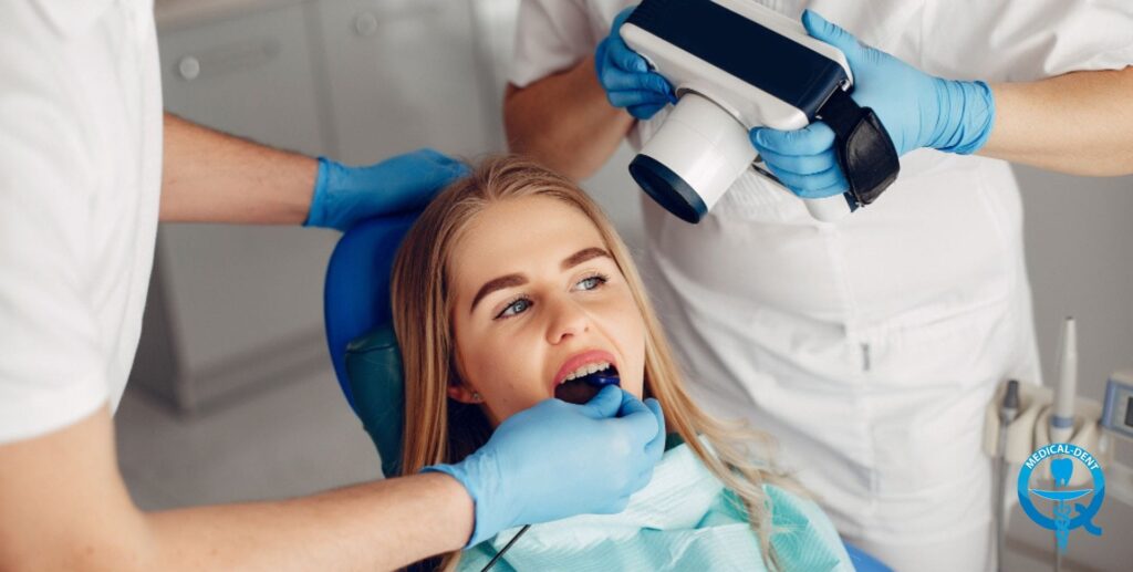 Kobieta w fotelu dentystycznym podczas zabiegu, z dwoma dentystami w rękawiczkach – jeden trzyma aparat rentgenowski, drugi używa narzędzia w jej ustach.